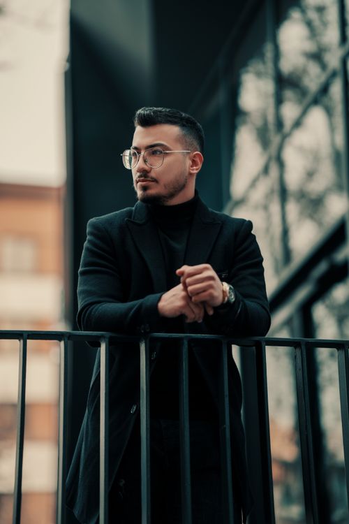 Free Elegant man with eyeglasses leaning on fencing in Tehran, wearing a black coat. Fashionable urban style. Stock Photo