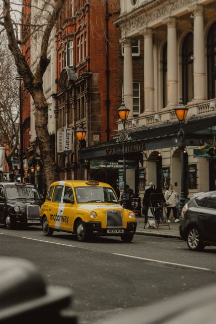 A yellow taxi driving on a bustling urban street in London, showcasing historic architecture.
