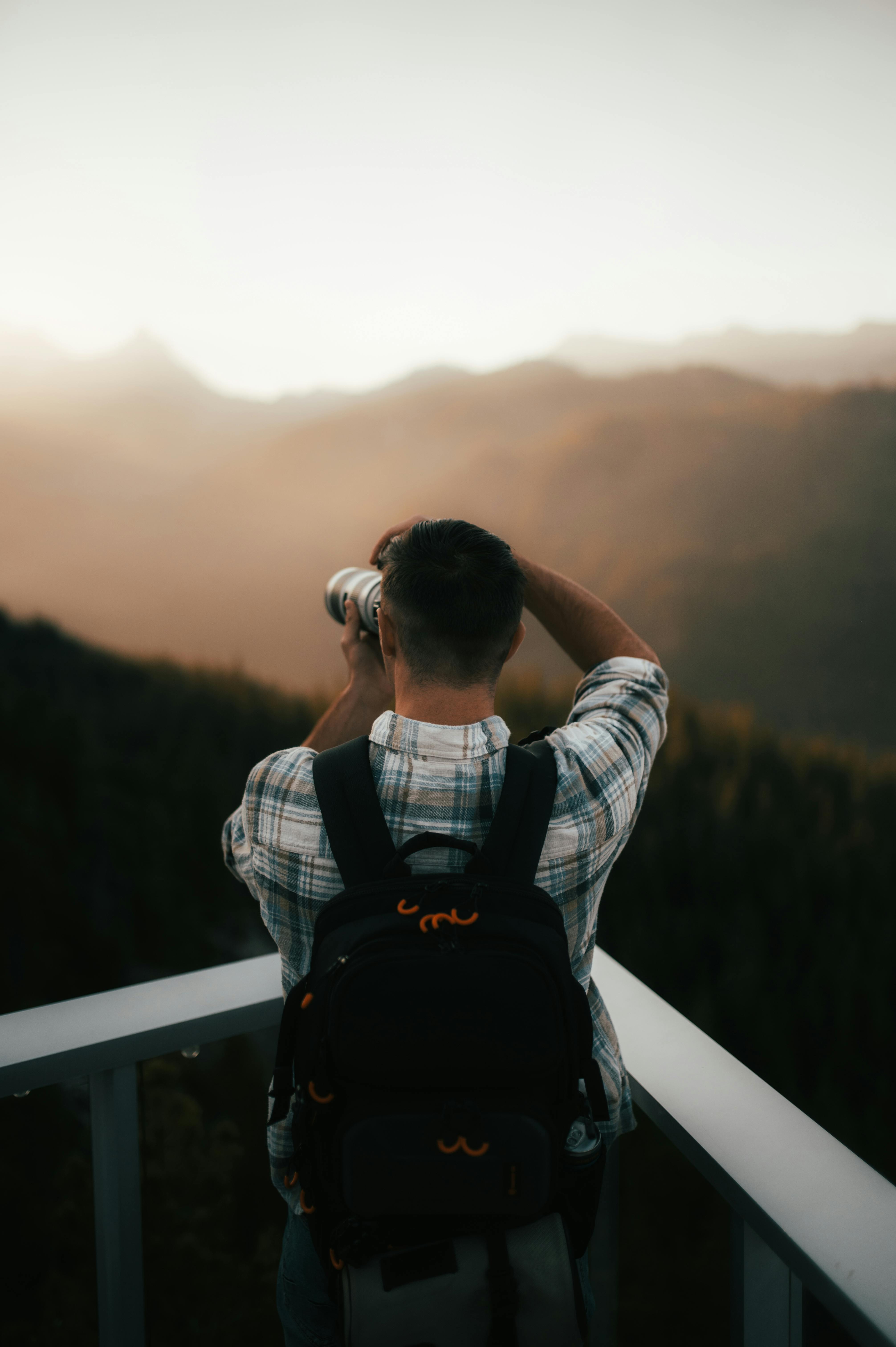 A photographer with a backpack takes pictures of a hazy mountain range during twilight from a balcony.