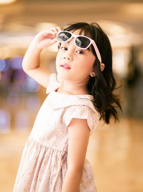Free Adorable young Asian girl in a pink dress posing confidently with sunglasses indoors. Stock Photo