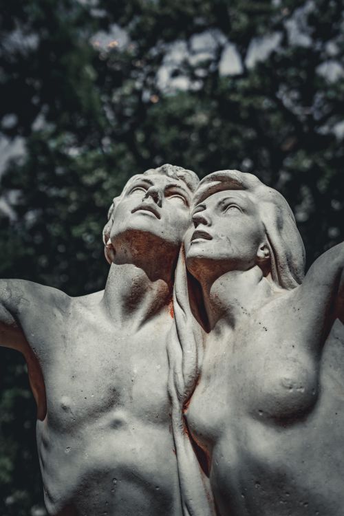 Free Close-up of a marble sculpture featuring a man and woman looking upwards in a lush outdoor setting. Stock Photo