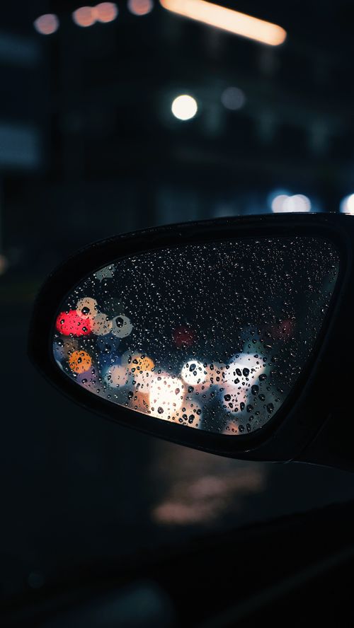 Free A close-up of raindrops on a car mirror reflecting blurred city lights at night. Stock Photo
