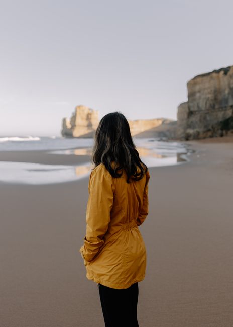 A woman in a yellow jacket stands on Anglesea beach with cliffs in view.