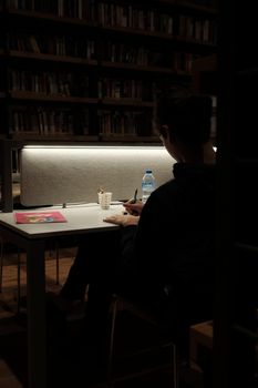 A person sits in a dimly lit library writing notes, surrounded by books.
