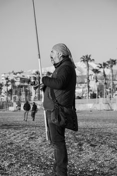 A fisherman stands on a pebble beach holding a fishing rod in black and white.
