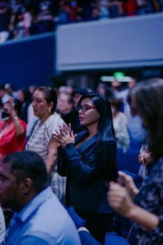 A diverse audience stands in anticipation at an indoor event.