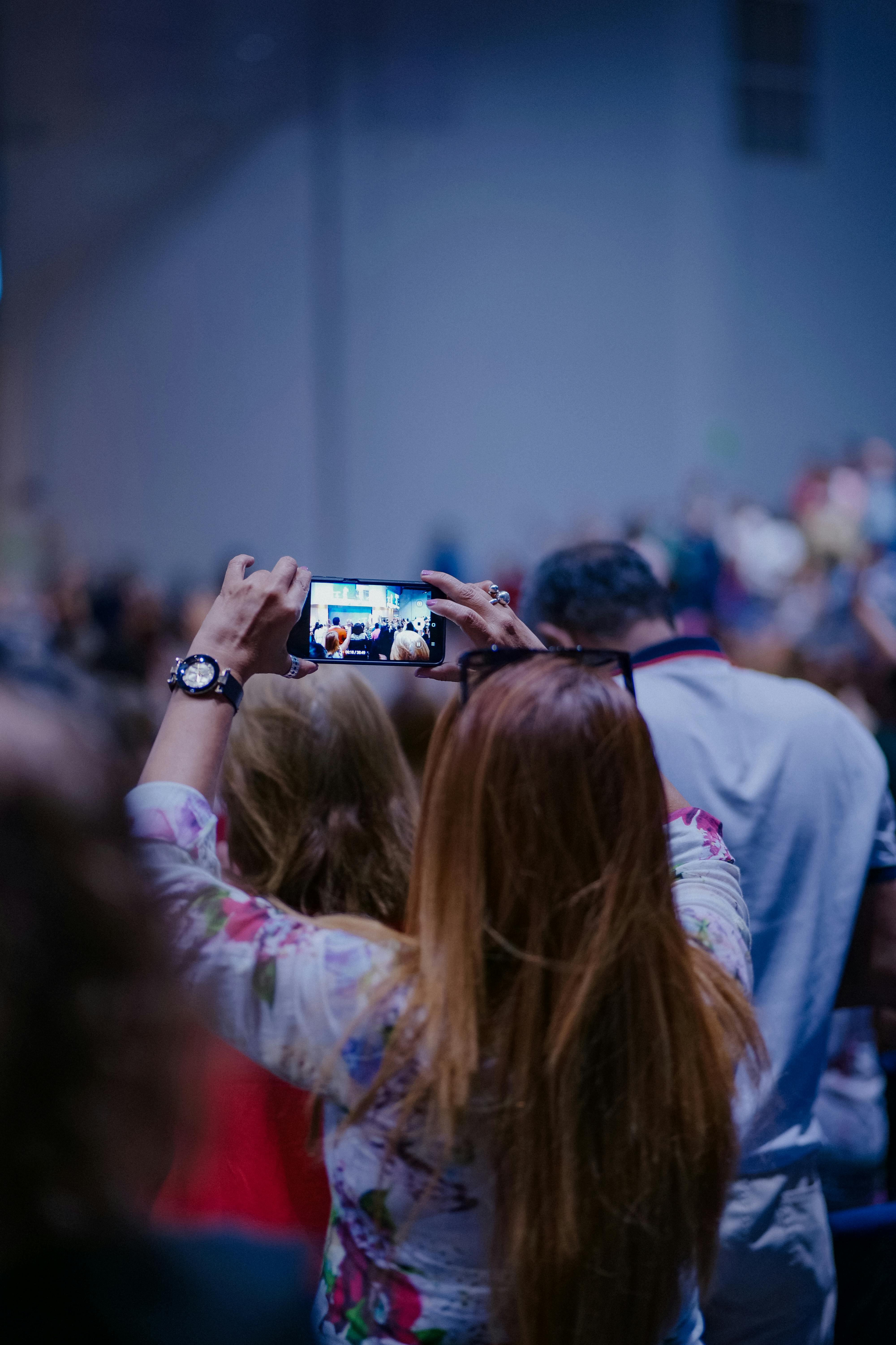 A woman records a live event with her smartphone amid a crowd, focusing on the stage.