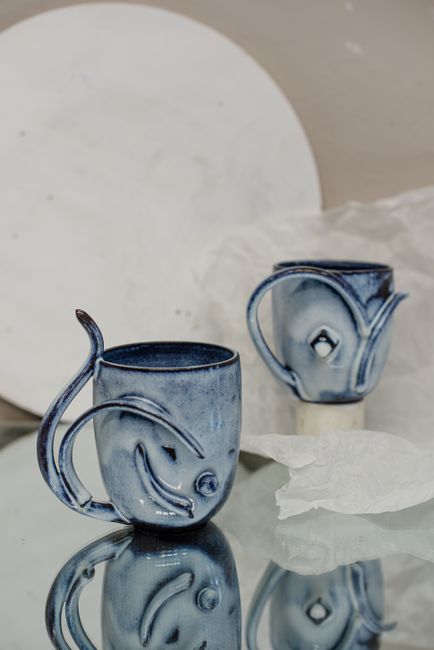 Handcrafted blue ceramic mugs with artistic designs reflecting on glass surface.