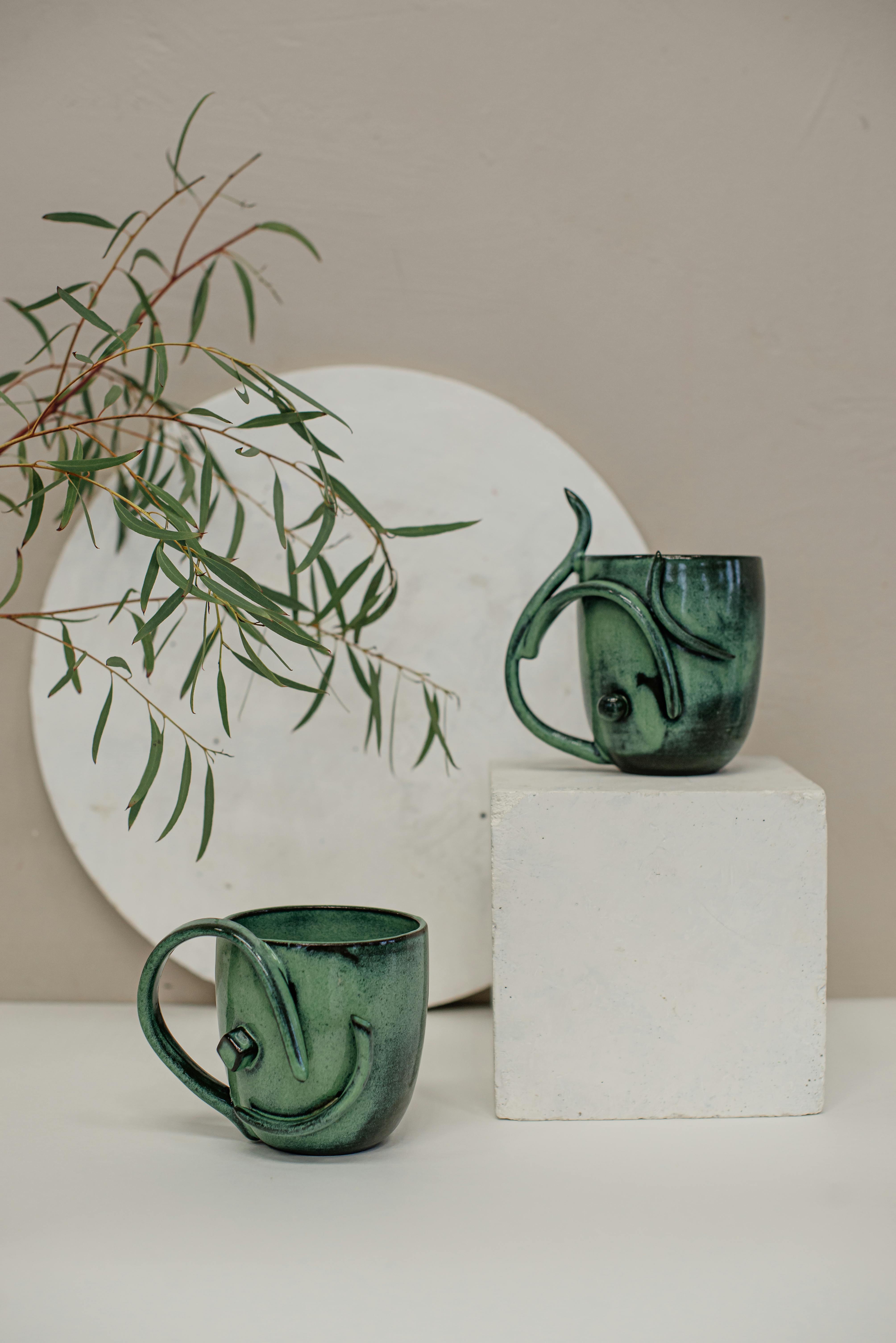 Elegant green ceramic mugs displayed in a minimalist still life arrangement with natural elements.