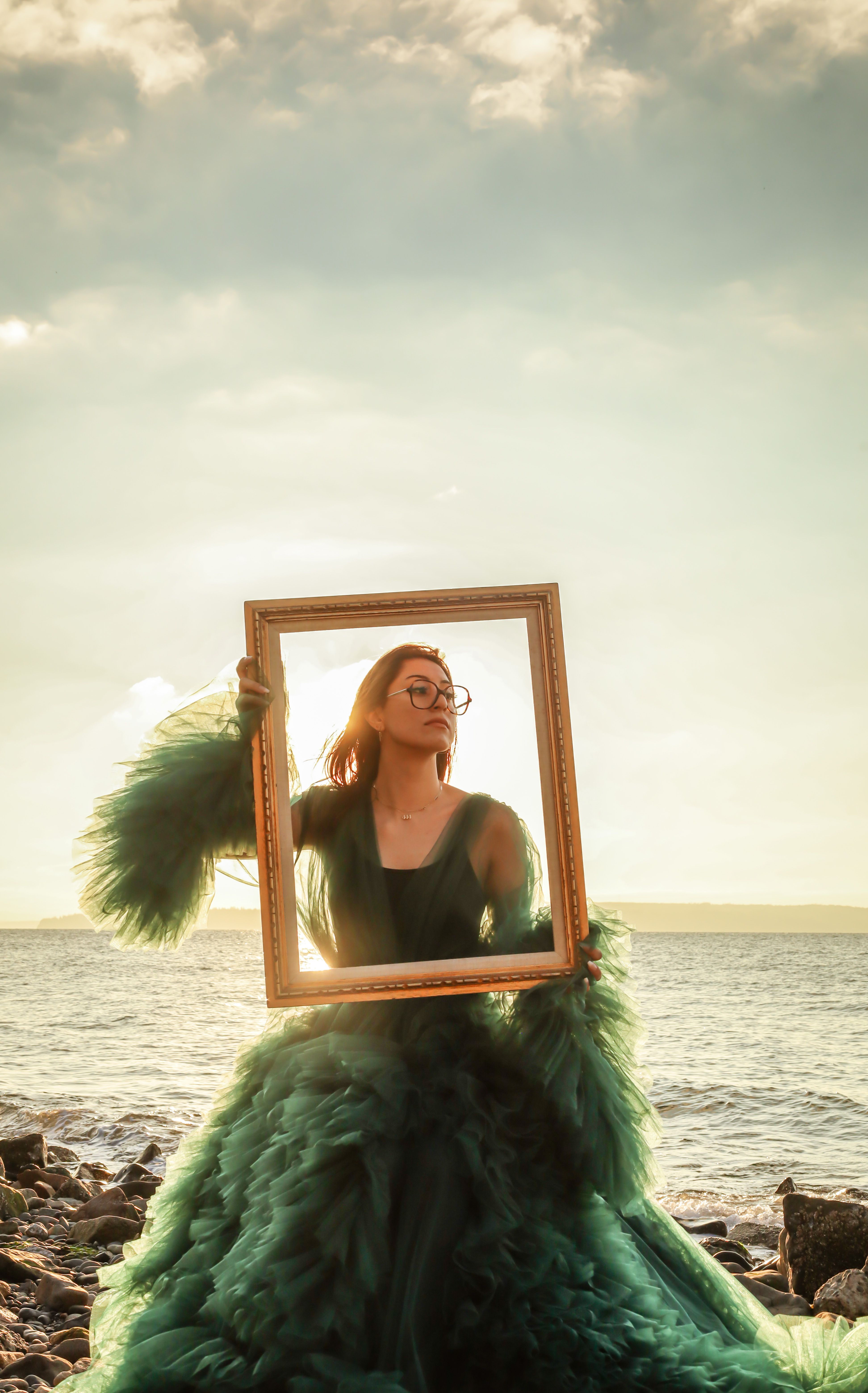 Elegant portrait of a woman in a green dress holding a frame by the sea during sunset.