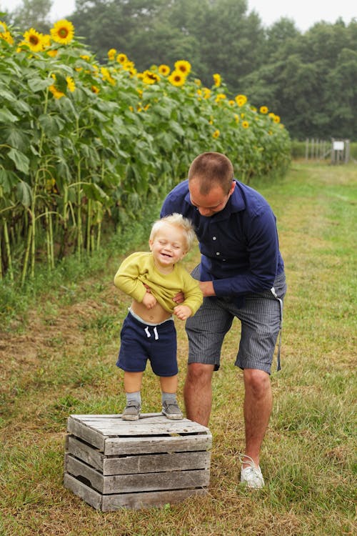 Free Man Holding Smiling Child Standing on Brown Wooden Crate Near Sunflowers Stock Photo