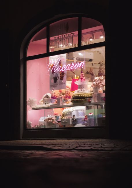 Charming night view of a macaron shop window with vibrant pink decor in Prague.