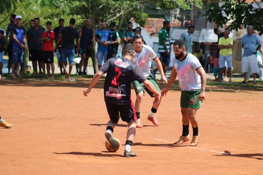 A lively amateur football game with players in action and an engaged crowd watching on a sunny day.