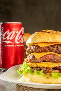 Mouthwatering cheeseburger stacked with ingredients next to a Coca-Cola can on a plate.