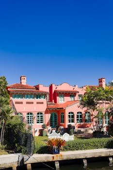 Elegant pink mansion in Fort Lauderdale with lush greenery and vibrant blue sky, showcasing luxury architecture.
