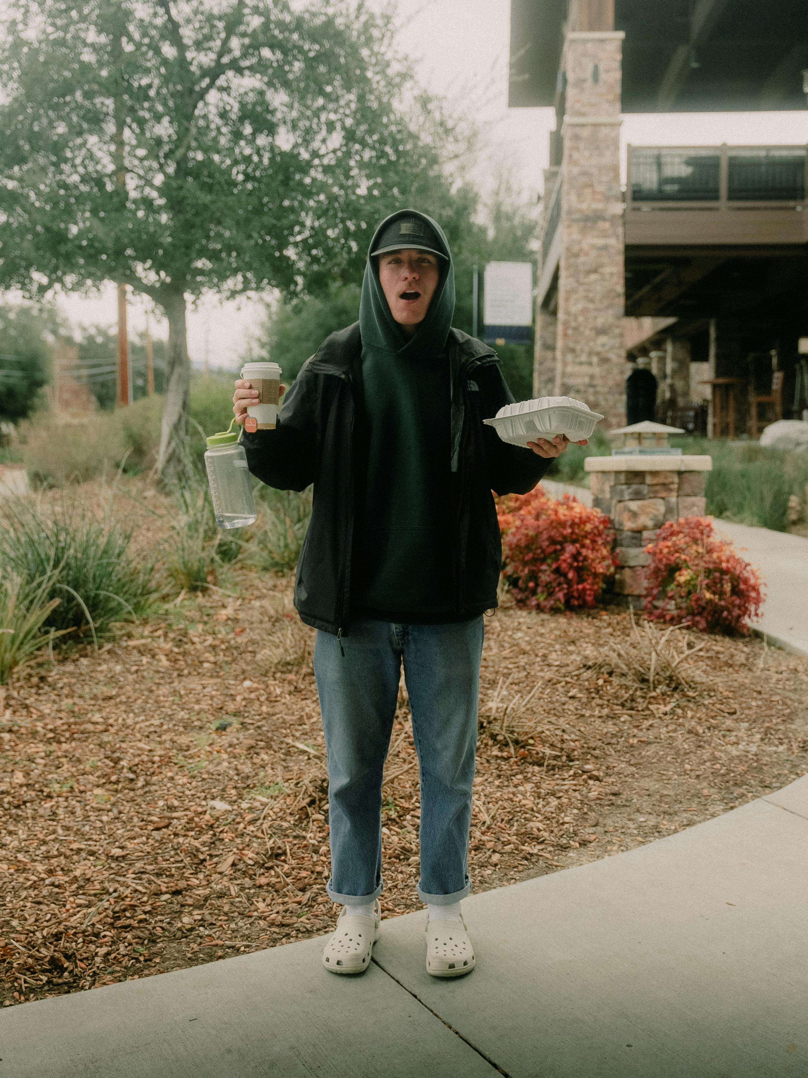 Casual individual outdoors holding a coffee cup and takeout in a natural setting.