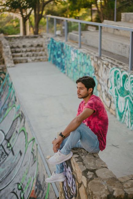 A young man sitting on a wall in a graffiti-filled skatepark, wearing casual fashion.