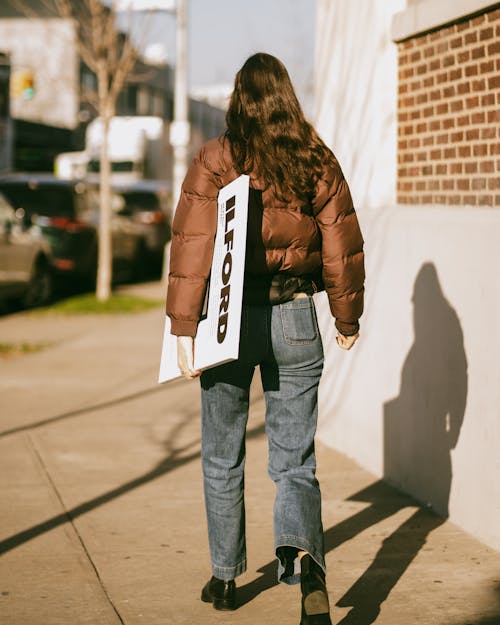 A Back View of a Woman in Brown Puffer Jacket Walking on the Street while Holding a Placard