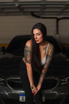 Stylish tattooed woman poses confidently in a São Paulo garage with a sleek car backdrop.
