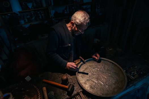 An artisan works meticulously on a metal plate in a dimly lit workshop in Denizli, Türkiye.