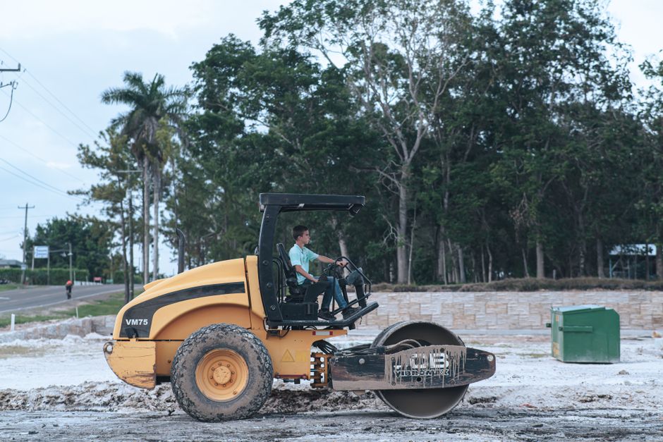 Teenager operates a road roller on a rural construction site, bordered by trees.