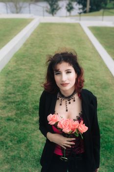 Caucasian woman with beaded necklace holding pink roses outdoors.