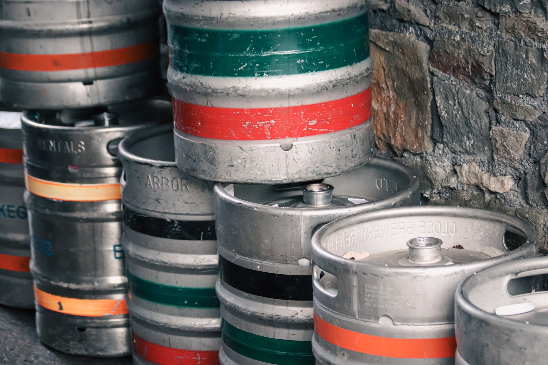 A collection of metal beer kegs stacked against a stone wall in a brewery storage area.