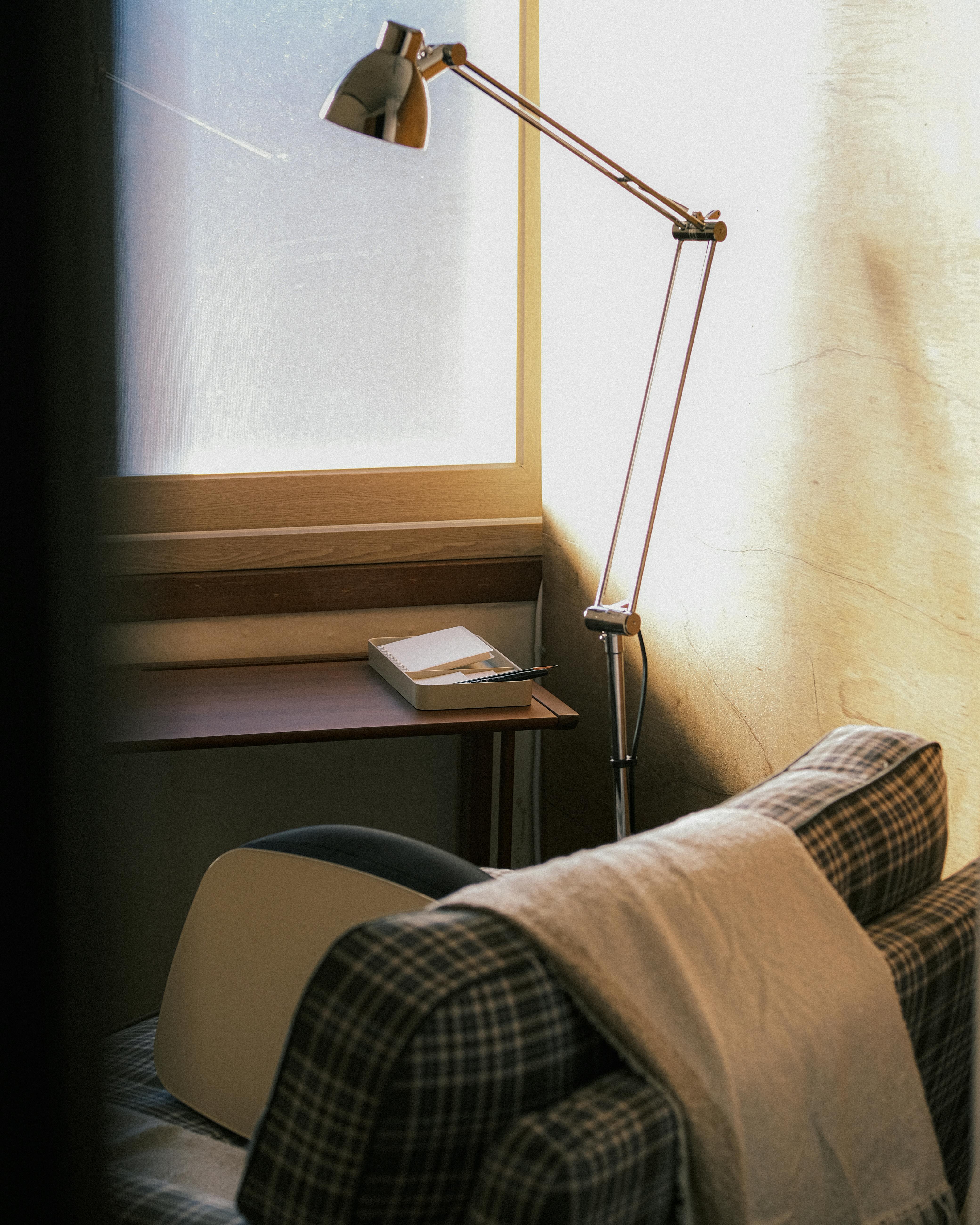 Free A cozy reading nook with a plaid chair, blanket, and a lamp by a window for serene relaxation. Stock Photo