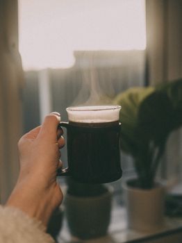 A hand holding a steaming mug of coffee by the window, capturing warmth.