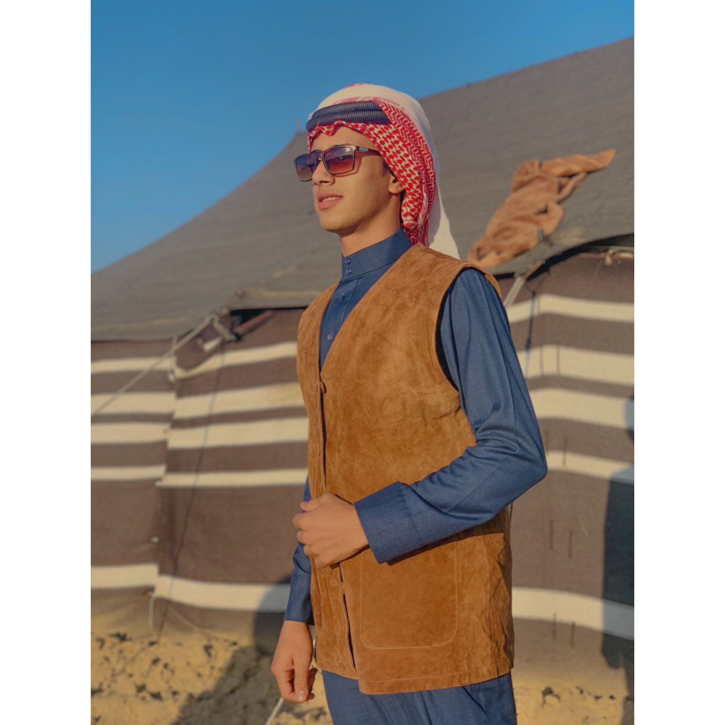 Portrait of a young man wearing traditional Middle Eastern attire standing outside a tent in sunlight.