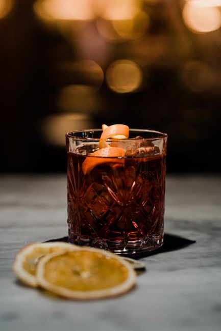 A small glass of dark amber amaro over a single large ice cube, garnished with an orange twist, next to the bottle and its herbal botanicals.