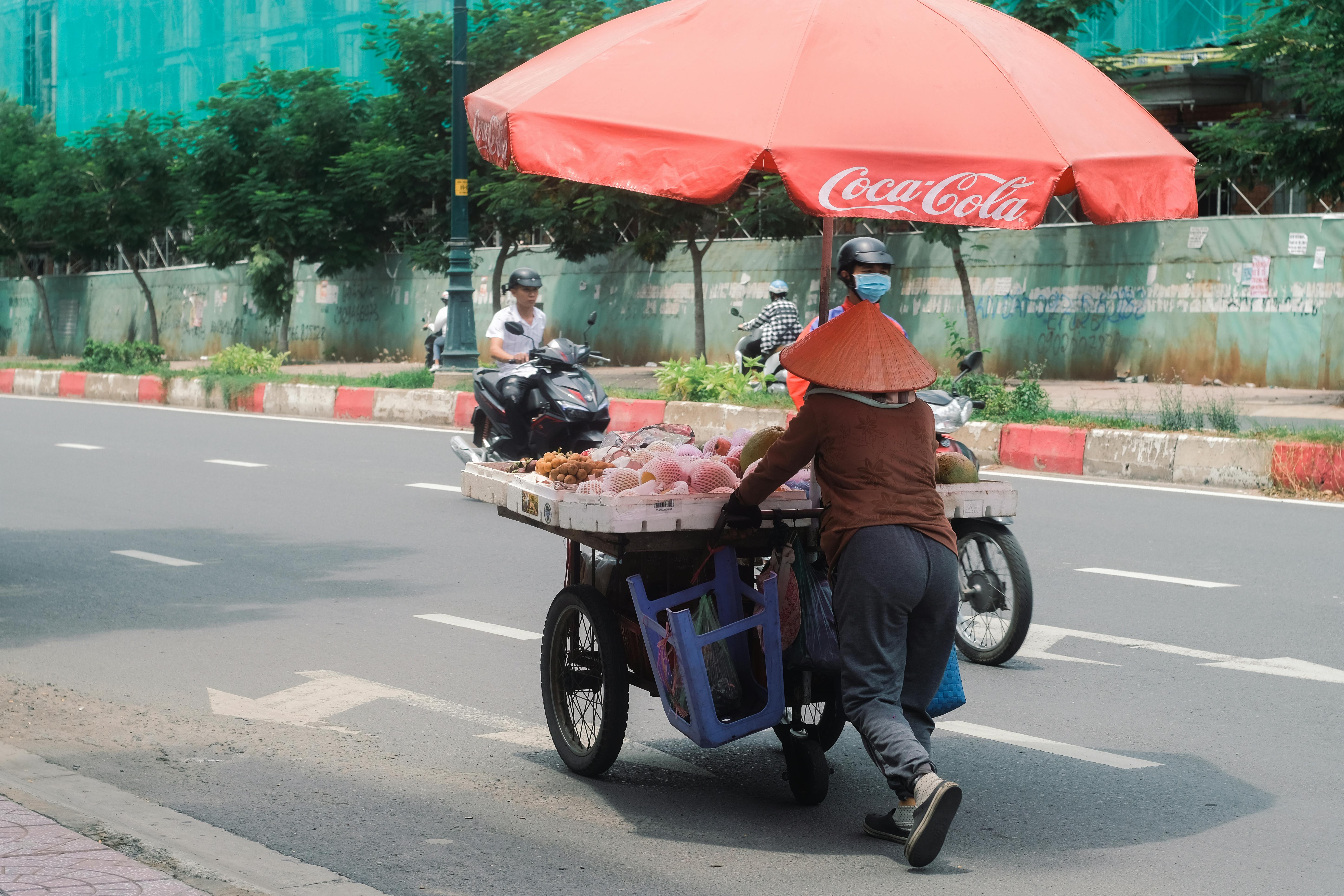 A street vendor pushes a fruit cart with a red umbrella on a city road.