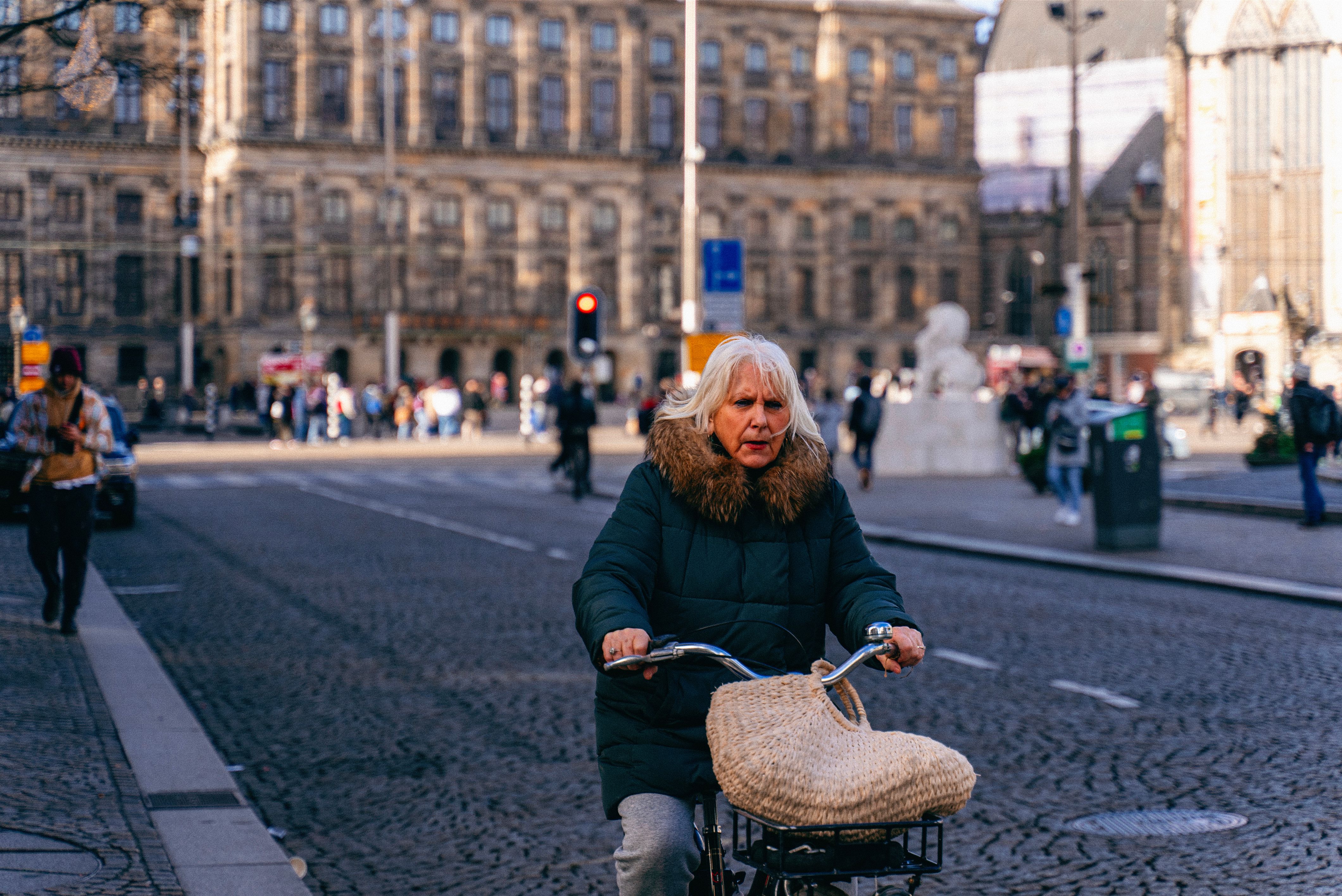 An elderly woman with gray hair rides a bicycle wearing a puffer jacket on a cobblestone street in an urban setting.