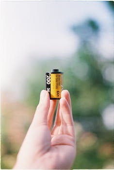 A close-up of a hand holding a vintage 35mm film roll with blurred outdoor background.