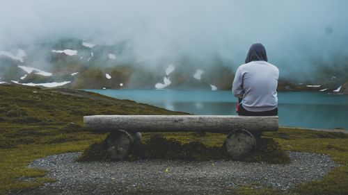 Free A person sits alone on a wooden bench by a misty lake, surrounded by mountains. Stock Photo