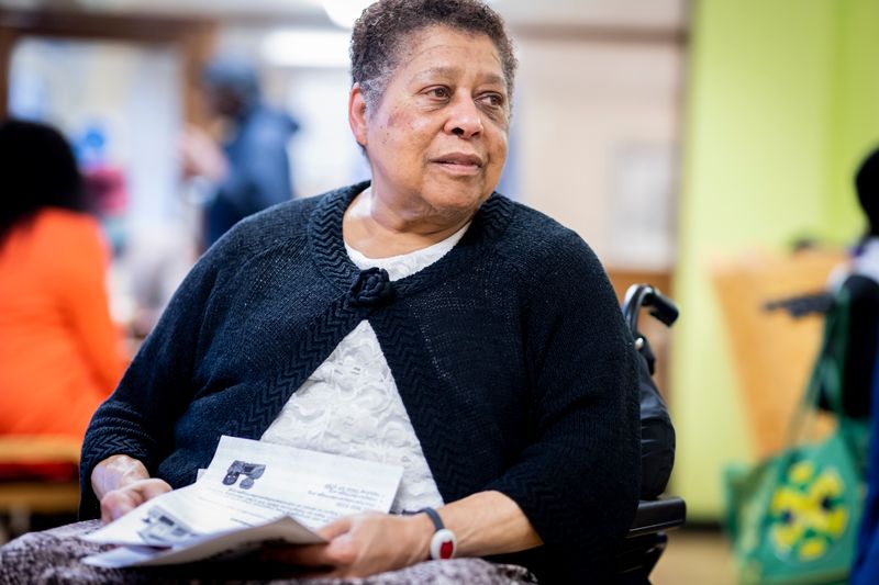 An older Black woman in a wheelchair holds a printed leaflet and paperwork, looking past the camera in a day room with other people behind her.