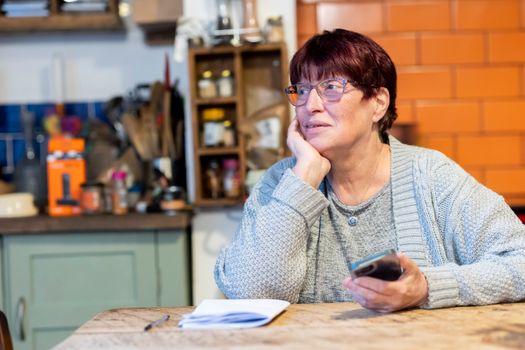 Senior woman in a cardigan pondering while holding a smartphone at a kitchen table.
