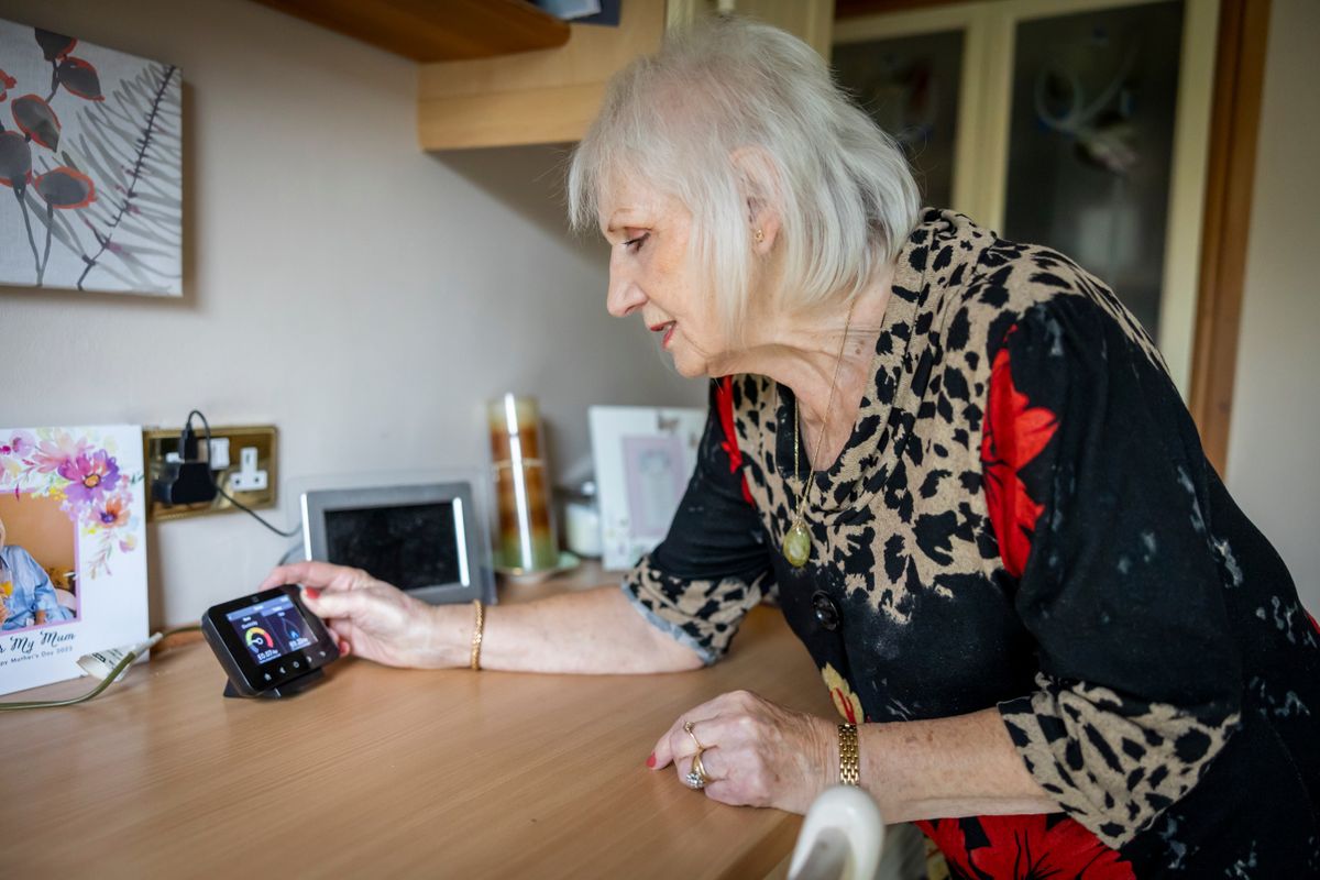 Mujer mayor de pelo blanco consultando la pantalla de un pequeño monitor de consumo energético apoyado sobre la encimera de su cocina