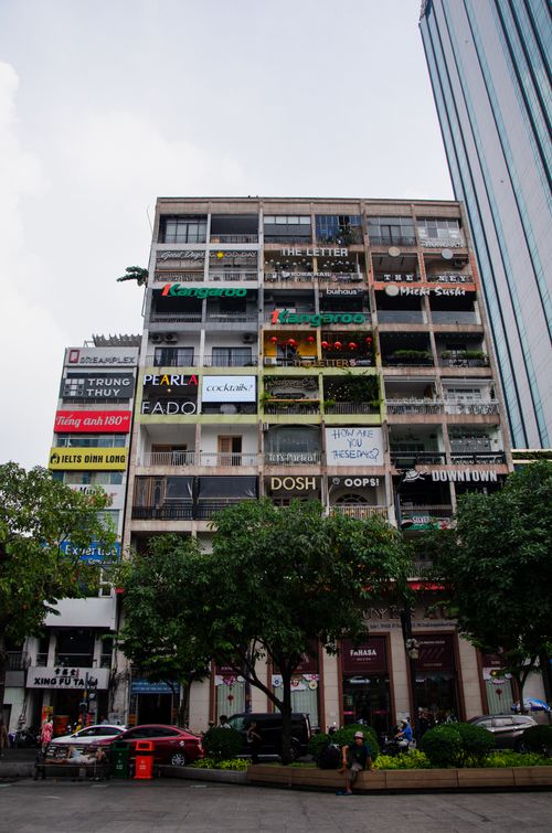 Free Modern urban high-rise building with diverse shop signs, trees, and bustling street view. Stock Photo