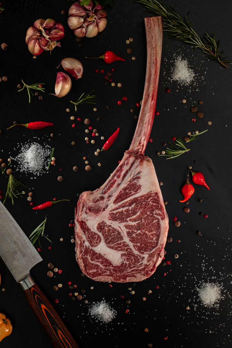 Close-up of raw tomahawk steak on a dark surface with herbs and spices.