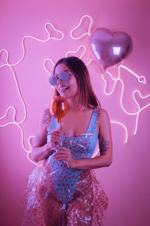 young and beautiful girl, in a blue bodysuit and a transparent red dotted dress holding a giant plastic lollipop, in front of a pink background with heart-shaped neons posing joyfully on a...