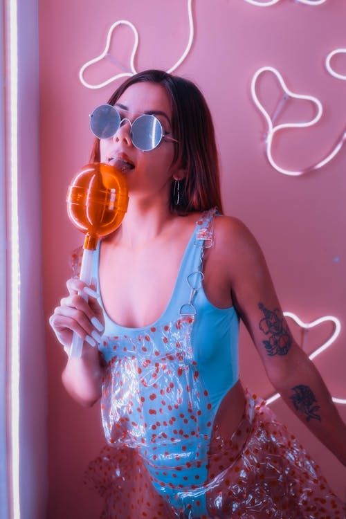 young and beautiful girl, in a blue bodysuit and a transparent red dotted dress holding a giant plastic lollipop, in front of a pink background with heart-shaped neons posing joyfully on a...