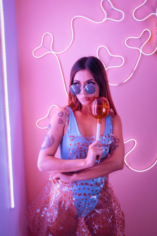 young and beautiful girl, in a blue bodysuit and a transparent red dotted dress holding a giant plastic lollipop, in front of a pink background with heart-shaped neons posing joyfully on a...