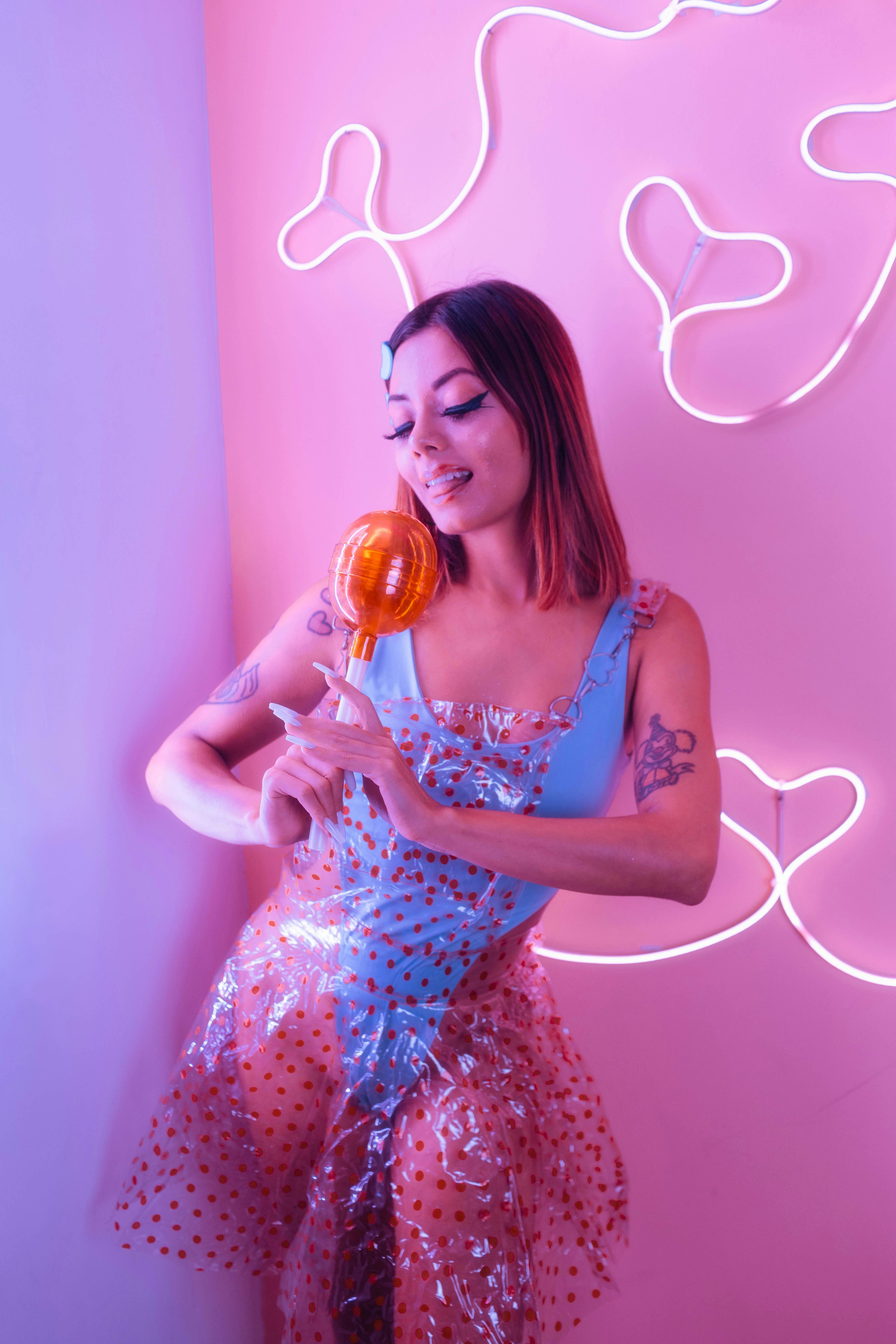 young and beautiful girl in a blue bodysuit and a transparent red dotted dress holding a giant plastic lollipop in front of a pink background with heart shaped neons posing joyfully on a