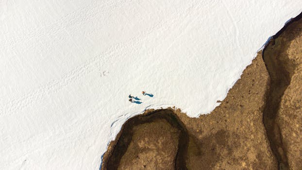 Aerial shot of hikers walking on snow beside a rugged brown landscape.