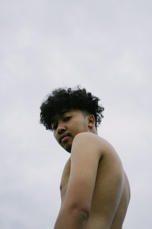 Free A young man with curly hair, shirtless, posing against a cloudy sky. Stock Photo