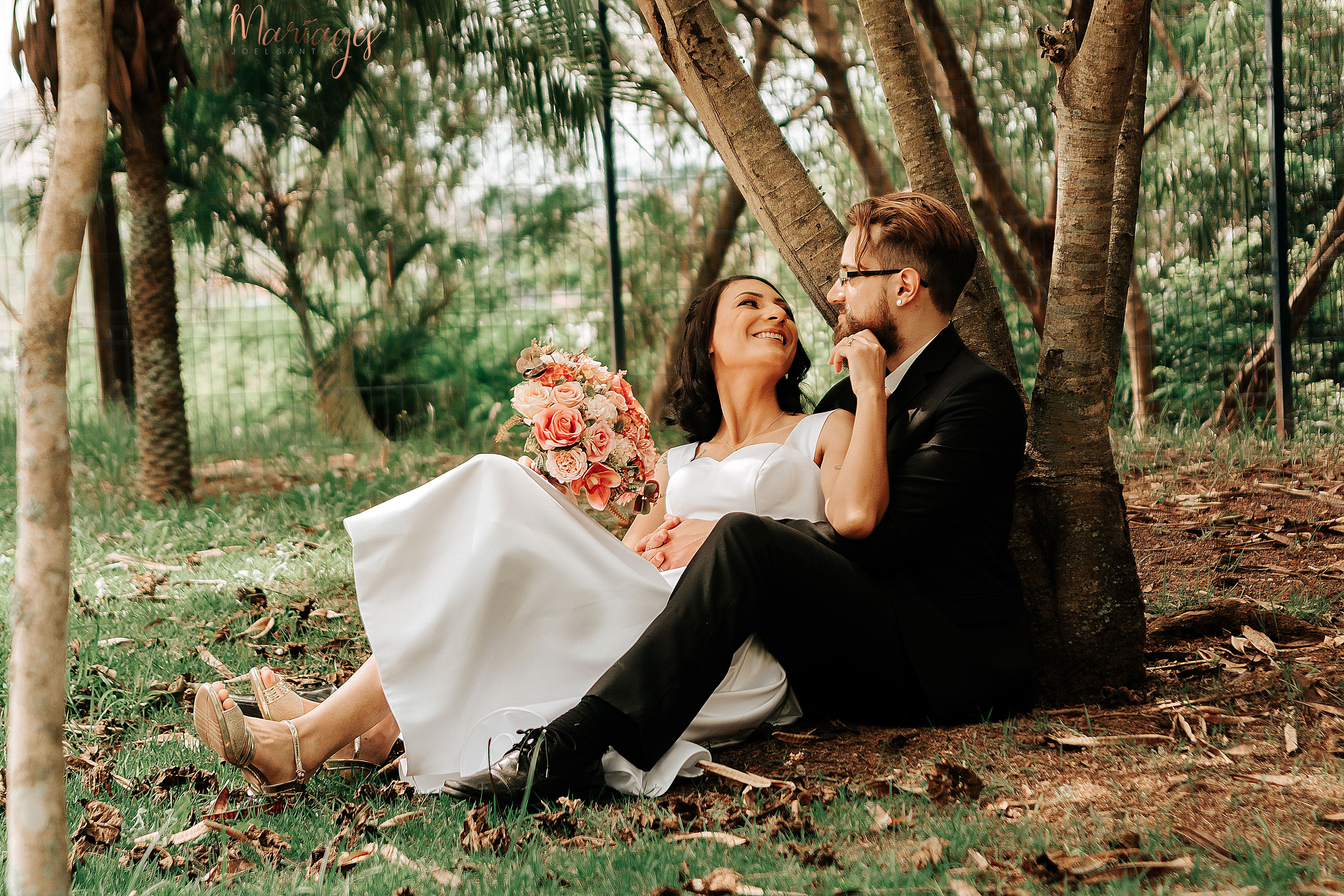 Bride and groom sitting under a tree, sharing a romantic moment with a bouquet of flowers in hand.