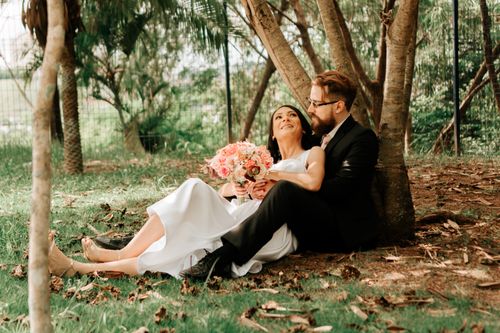 Free A joyful bride and groom sitting under trees, embracing each other outdoors. Stock Photo