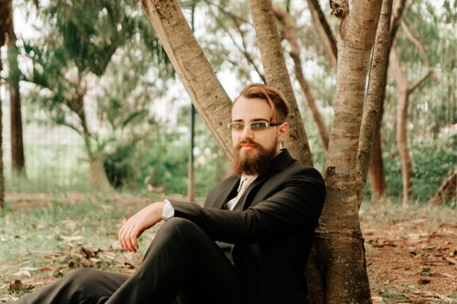 Free A well-dressed man with a beard sits pensively under a tree in a serene park setting. Stock Photo