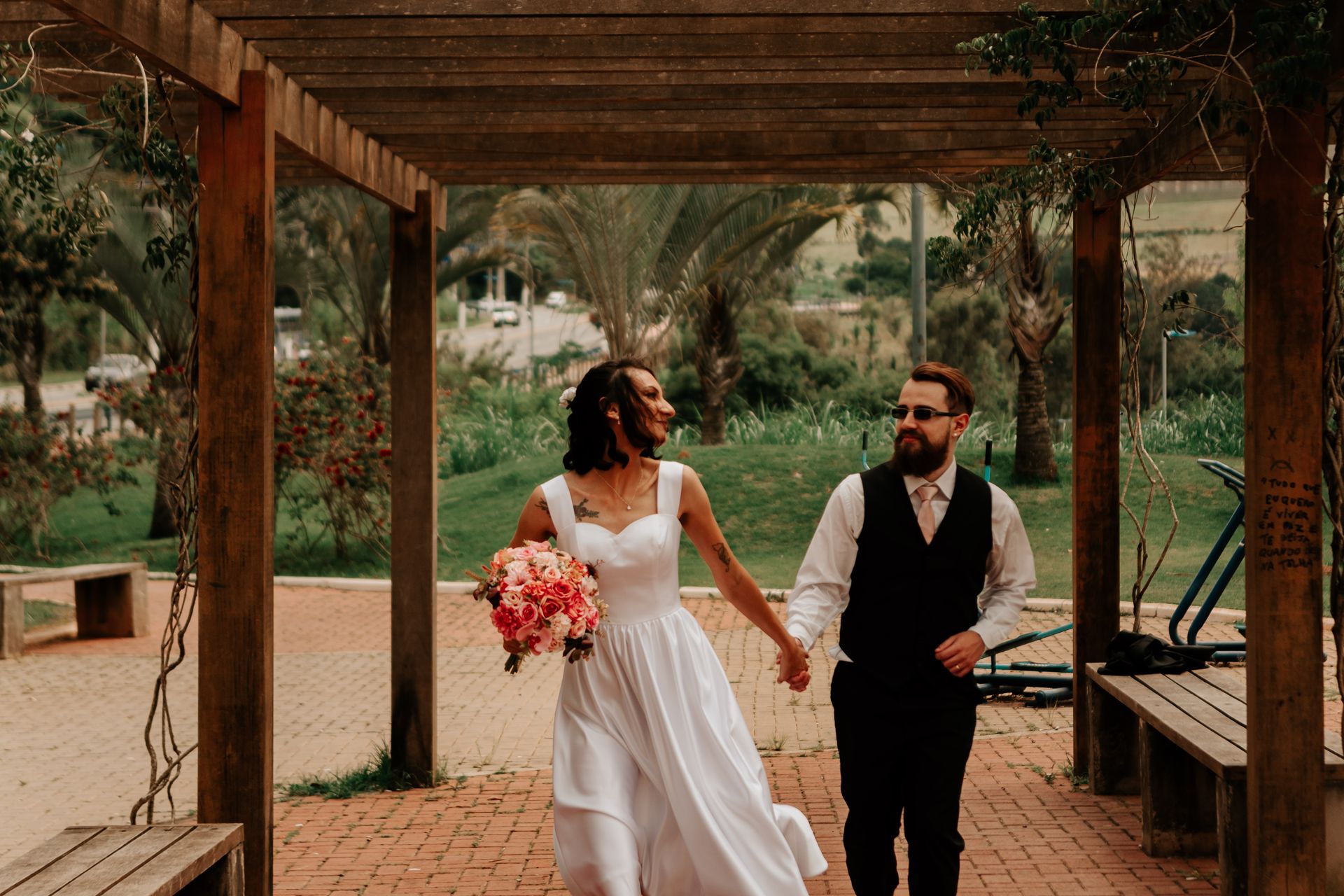 A happy couple in wedding attire walking hand-in-hand under a wooden pergola on their special day.